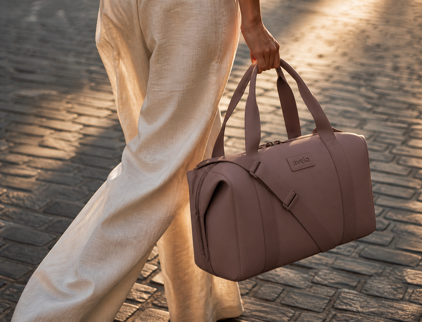 Woman carrying an Avela mauve neoprene carryall on a cobblestone street in golden hour light